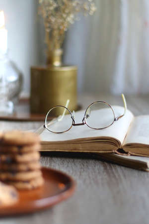 Stack of chocolate chip cookies, open book and reading glasses, lit candle and vase with flowers on the table. Hygge at home. Selective focus.の写真素材