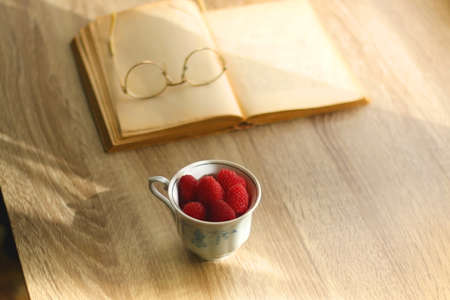 Porcelain cup filled with raspberries, open book, reading glasses and vase with flowers on the table. Selective focus.の写真素材
