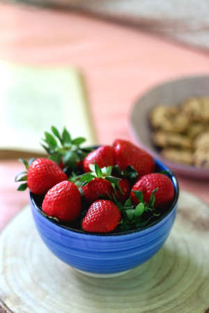 Bowl of chocolate chip cookies, bowl of fresh strawberries, open book and reading glasses on the bed. Hygge at home. Selective focus.の写真素材