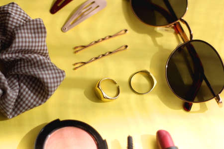 Retro sunglasses, gold rings, various hair accessories and make up products on vibrant yellow background. Selective focus.の写真素材