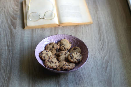 Bowl of chocolate chip cookies, open book and reading glasses on the table. Selective focus.の写真素材