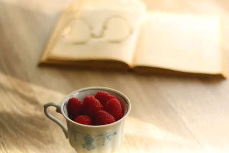Porcelain cup filled with raspberries, open book, reading glasses and vase with flowers on the table. Selective focus.の写真素材