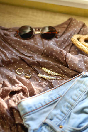 Brown polka dot blouse, vintage mom jeans, straw bag, round sunglasses, gold rings and pearl hair clips. Fashionable outfit on wooden background. Selective focus.の写真素材