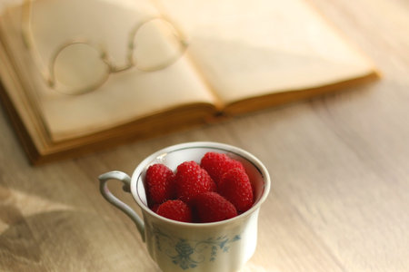 Porcelain cup filled with raspberries, open book, reading glasses and vase with flowers on the...