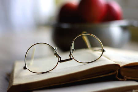 Silver bowl filled with red apples, open book and reading glasses on the table. Selective focus.の写真素材