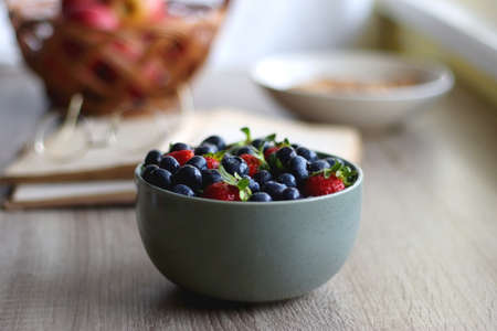 Bowl of strawberries and blueberries, open books with reading glasses, plate of chocolate chip cookies and basket of apples one the table. Selective focus.の写真素材