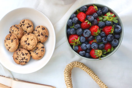 Picninc blanket with straw bag, bowl of strawberries and blueberries, bowl of chocolate chip cookies, books, sunglasses and basket of apples in the garden. Top view.の写真素材