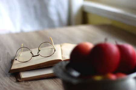 Silver bowl filled with red apples, open book and reading glasses on the table. Selective focus.の写真素材