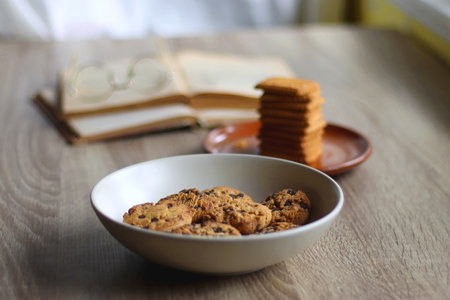 Bowl of chocolate chip cookies, plate of biscuits, open books and reading glasses on the table....