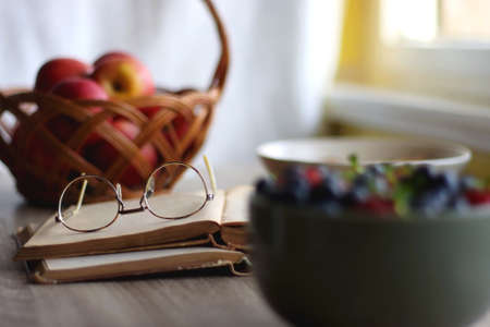 Bowl of strawberries and blueberries, open books with reading glasses, plate of chocolate chip cookies and basket of apples one the table. Selective focus.の写真素材
