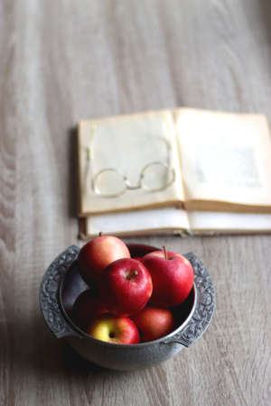 Silver bowl filled with red apples, open book and reading glasses on the table. Selective focus.の写真素材