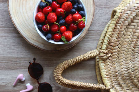 Round straw bag, brown sunglasses, pink earphones and bowl of berries on wooden table. Flat lay.の写真素材