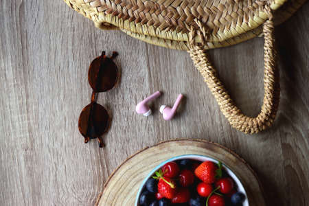 Round straw bag, brown sunglasses, pink earphones and bowl of berries on wooden table. Flat lay.の写真素材