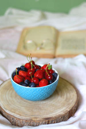 Bowl of strawberries, cherries and blueberries, open book and reading glasses on the bed. Selective focus.の写真素材