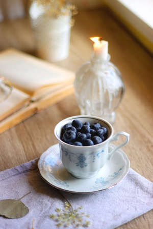 Vintage porcelain cup filled with fresh blueberries, pressed flowers, open book, reading glasses and...