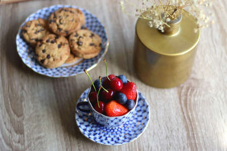 Plate of chocolate chip cookies, cup filled with strawberries, blueberries and cherries, open book...