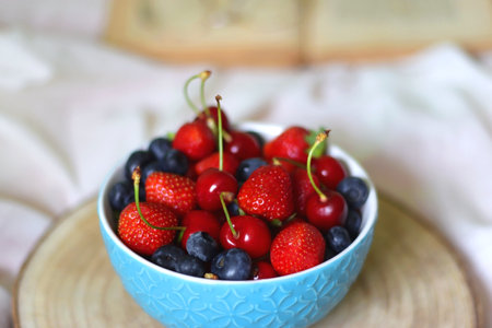 Bowl of strawberries, cherries and blueberries, open book and reading glasses on the bed. Selective focus.の写真素材