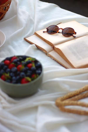 Picninc blanket with straw bag, bowl of strawberries and blueberries, bowl of chocolate chip cookies, books, sunglasses and basket of apples in the garden. Selective focus.の写真素材