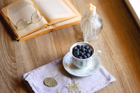 Vintage porcelain cup filled with fresh blueberries, pressed flowers, open book, reading glasses and...