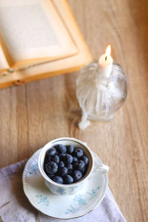 Vintage porcelain cup filled with fresh blueberries, pressed flowers, open book, reading glasses and lit candle on the table. Hygge at home. Selective focus.の写真素材