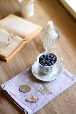 Vintage porcelain cup filled with fresh blueberries, pressed flowers, open book, reading glasses and lit candle on the table. Hygge at home. Selective focus.の写真素材