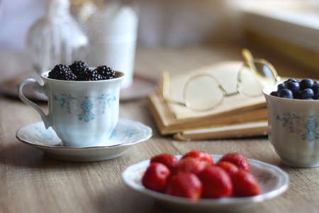 Vintage tea set filled with blueberries, blackberries and starwberries, open book, reading glasses...