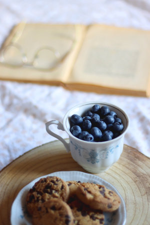 Vintage cup with fresh blueberries, plate of chocolate chip cookies, open book and reading glasses on a bed. Selective focus.の写真素材