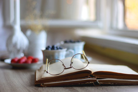 Vintage tea set filled with blueberries, blackberries and starwberries, open book, reading glasses and decorative candles on the table. Selective focus.の写真素材