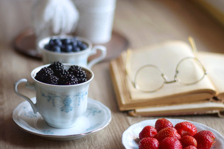 Vintage tea set filled with blueberries, blackberries and starwberries, open book, reading glasses and decorative candles on the table. Selective focus.の写真素材