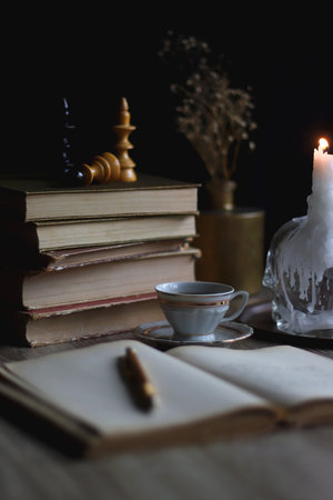 Stack of vintage books, cup of tea or coffee, lit candles, reading glasses and chess pieces on...