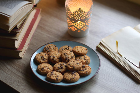 Plate of chocolate chip cookies, stack of vintage books, reading glasses, cup of tea or coffee, lit candle and fairy lights. Hygge at home. Selective focus.の写真素材