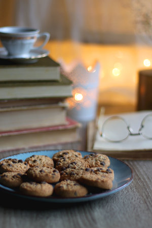 Plate of chocolate chip cookies, stack of vintage books, reading glasses, cup of tea or coffee, lit...