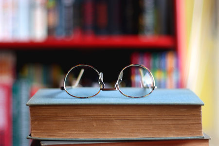 Stack of colorful hardcover books and reading glasses in front of a bookshelf. Selective focus.の写真素材