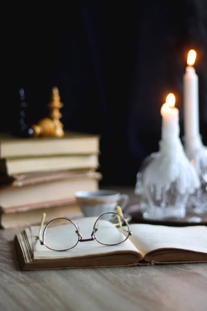 Stack of vintage books, cup of tea or coffee, lit candles, reading glasses and chess pieces on wooden table. Dark academia concept. Selective focus.の写真素材