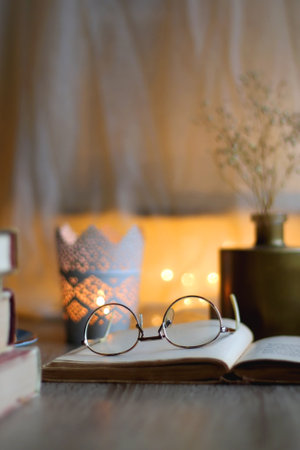 Plate of chocolate chip cookies, stack of vintage books, reading glasses, cup of tea or coffee, lit candle and fairy lights. Hygge at home. Selective focus.の写真素材