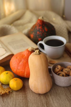 Cup of tea or coffee, seasonal spices, bowl of cookies, blanket, pumpkins, colorful leaves, books and tangerines on wooden table. Cozy hygge at home. Selective focus.の写真素材