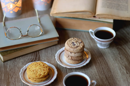 Plates with various cookies, cups of tea or coffee, vintage books, reading glasses and lit candles on the table. Selective focus.の写真素材