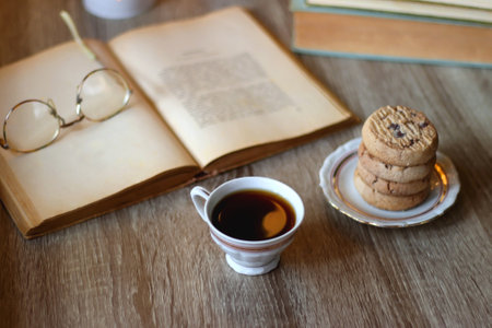 Plates with various cookies, cups of tea or coffee, vintage books, reading glasses and lit candles on the table. Selective focus.の写真素材