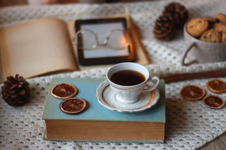 Cup of tea, books, tablet, bowl of cookies, various spices, pine cones and lit candles. Hygge at home. Selective focus.の写真素材