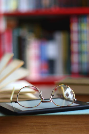 Stack of hardcover books, e-reader and reading glasses on the table. Bookshelf in the background....