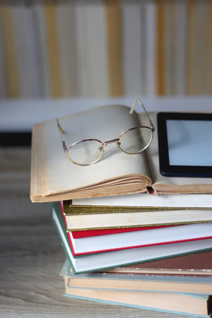 Stack of books on the table, reading glasses and e-reader on the table. Bookshelf in the background. Selective focus.の写真素材