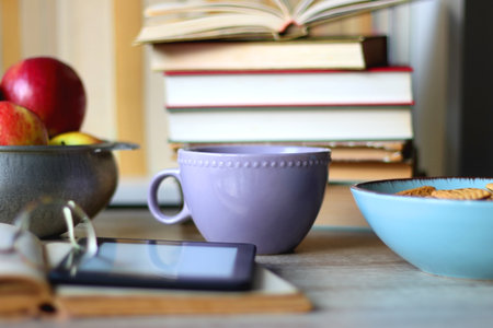 Stack of books, e-reader, reading glasses, bowl of biscuits, apples and cup of tea on the table. Bookshelf in the background. Selective focus.の写真素材
