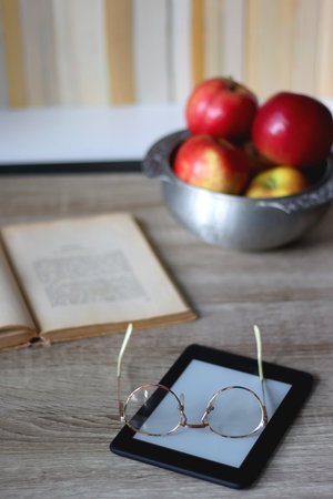 Open book, e-reader, reading glasses and bowl of apples on the table. Bookshelf in the background....