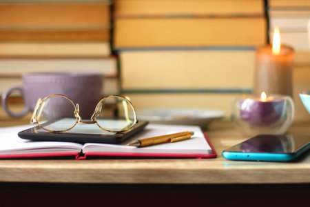 Notebook, pen, tablet, reading glasses, books, phone, candle, bowl of biscuits and cup of tea on the...