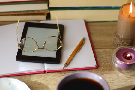Notebook, pen, tablet, reading glasses, books, phone, candle, bowl of biscuits and cup of tea on the table. Selective focus.の写真素材