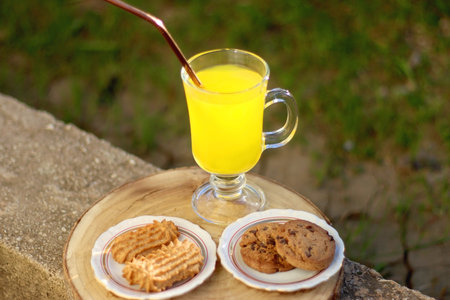 Glass of orange juice with reusable straw and two plates with cookies, served in a garden. Selective focus.の写真素材
