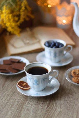 Cup of tea or coffee, plate of cookies, cup of blueberries, plate of chocolate, glass of juice, book, reading glasses, teapot, flowers and lit candles on the table.の写真素材