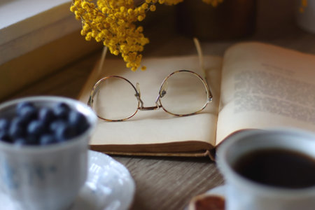 Cup of tea or coffee, plate of cookies, cup of blueberries, plate of chocolate, glass of juice, book, reading glasses, teapot, flowers and lit candles on the table.の写真素材