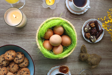 Easter eggs in the basket, bowl of cookies, chocolate pralines, Easter bunny figurine, cups of tea, glasses of juice, flowers and lit candles on the table. Top view.の写真素材
