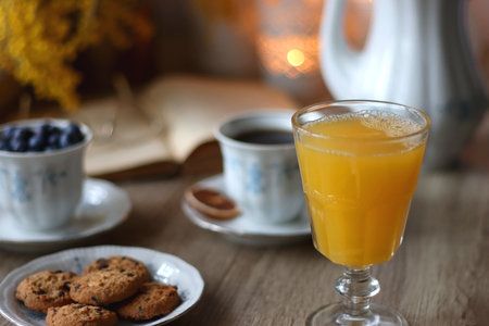 Cup of tea or coffee, plate of cookies, cup of blueberries, plate of chocolate, glass of juice, book, reading glasses, teapot, flowers and lit candles on the table.の写真素材